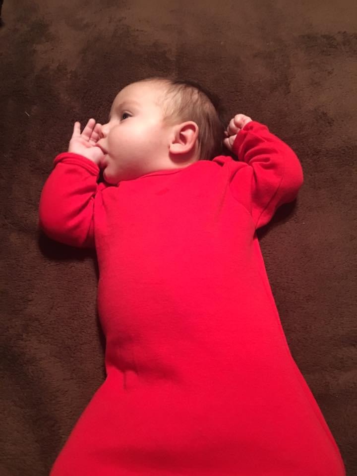 MacKenzy is lying on her back with her head turned to the right. Her arms are bent upward and her right thumb is in her mouth. She is wearing a long-sleeved, red cotton nightgown.