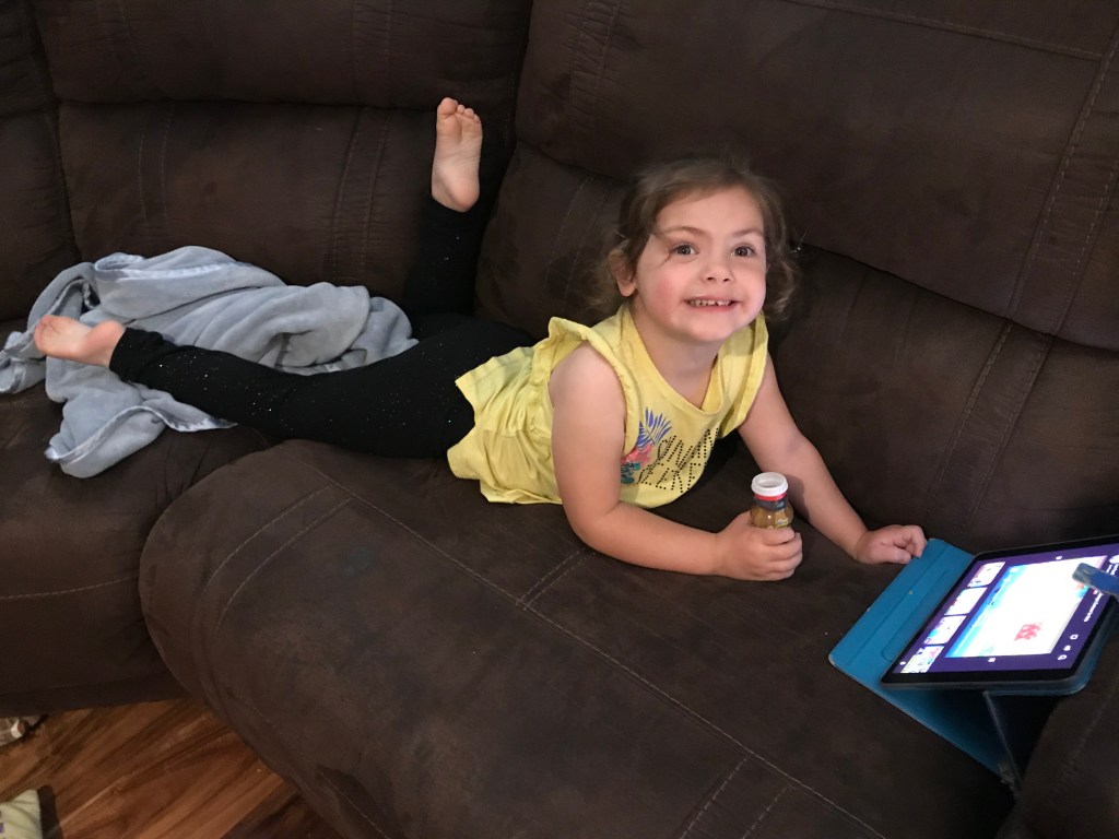 MacKenzy is lying on her stomach propped up on her elbows. She is on a couch watching an iPad with a Danimal yogurt drink in her right hand. She is wearing a yellow shirt and black legging pants.