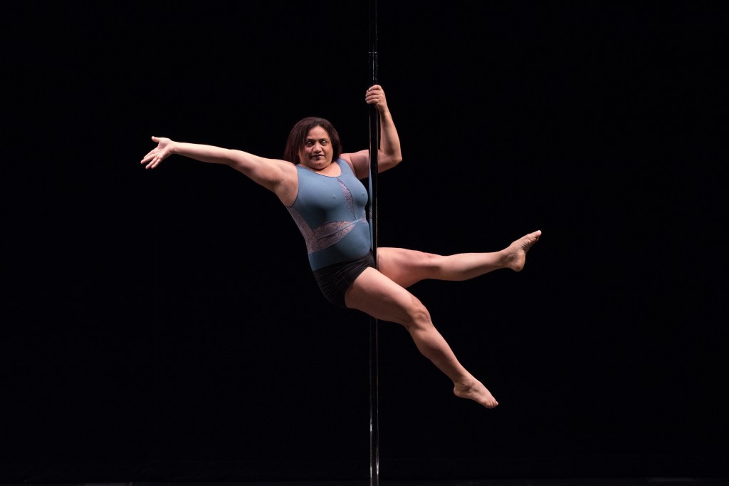 Side pose of LM halfway up the pole. Her right arm sweeps out behind her while her left hand and forearm are on the pole. Her thighs grip the pole and her legs are straight out in a V