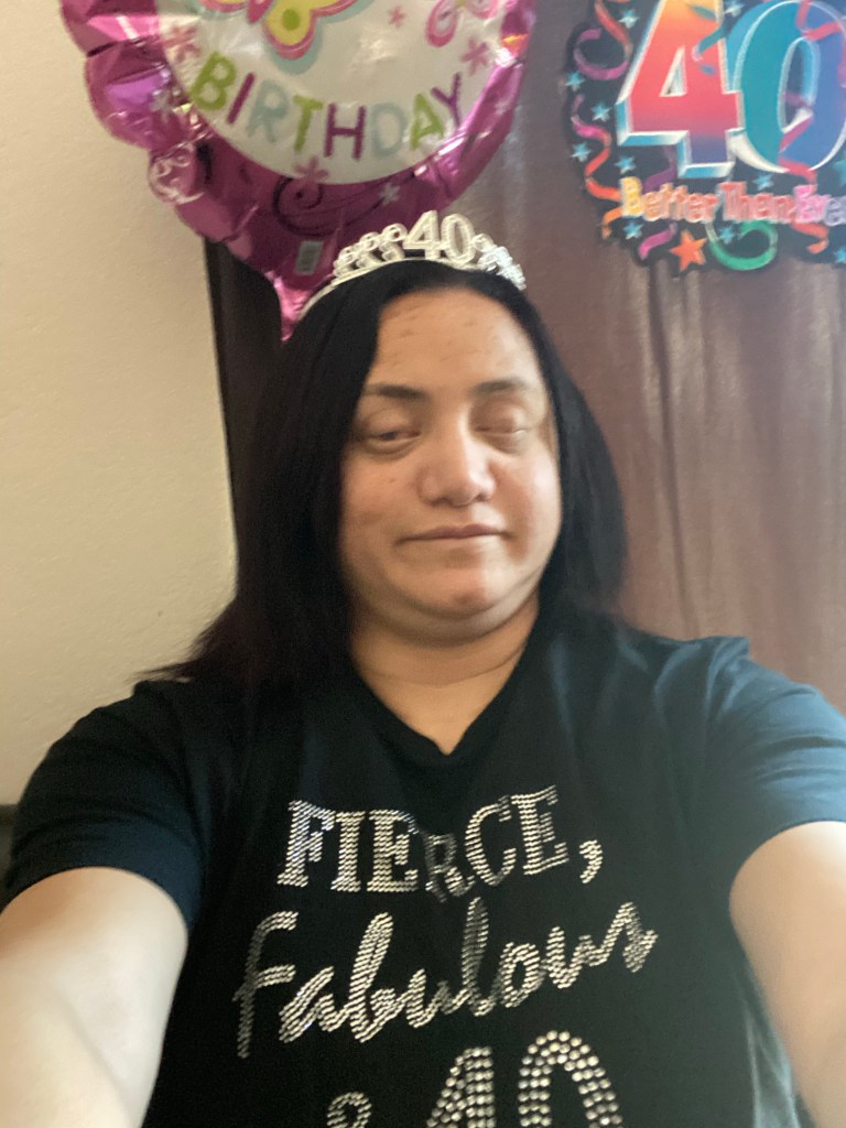 LM Sits in front of birthday decorations. She is wearing a crown with the number 40 in the center and a black shirt with rhinestones that say fierce, fabulous, and 40.