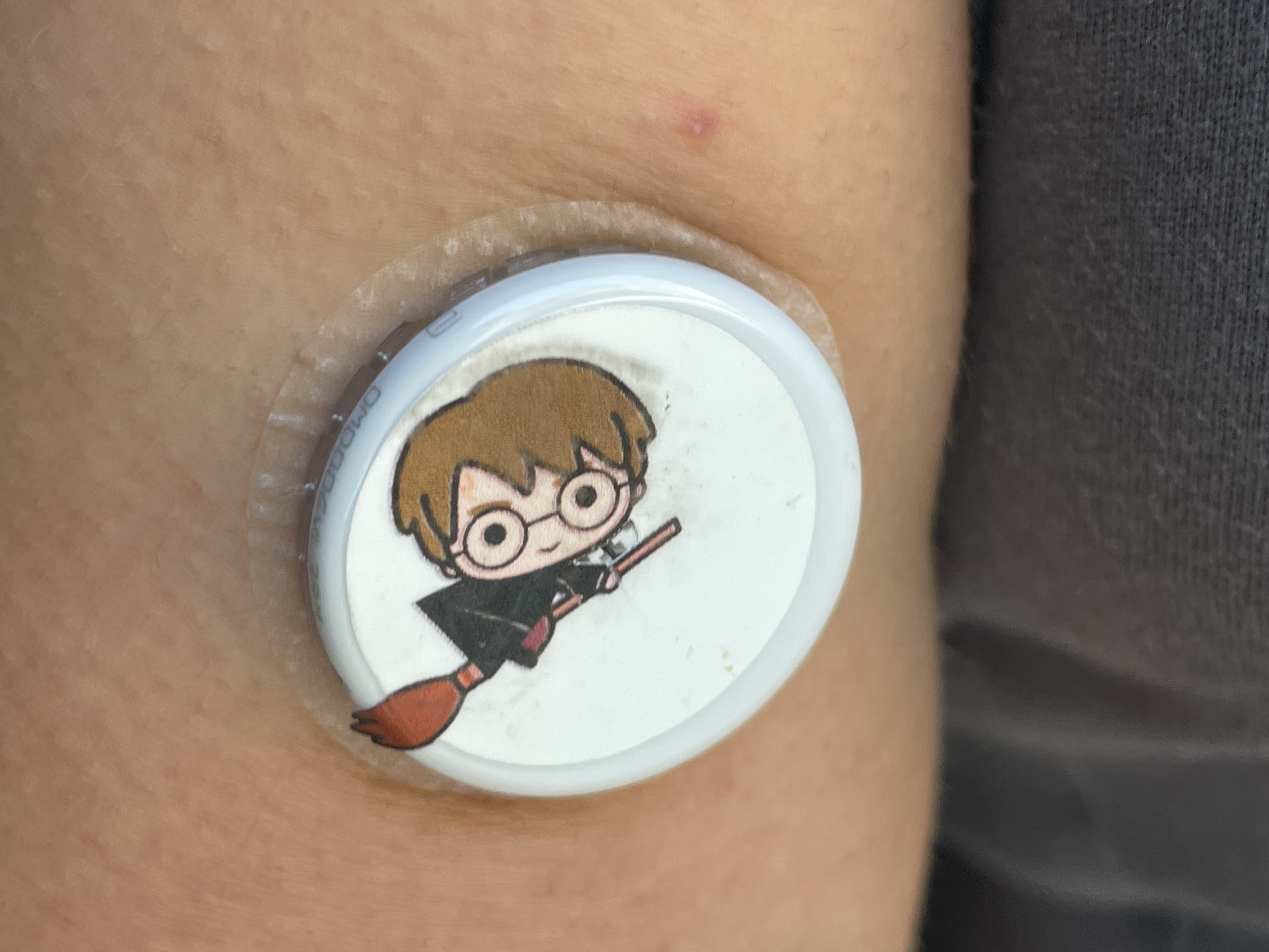 A FreeStyle Libre sensor applied to the back of a brown arm. It is covered with a sticker depicting a Harry Potter figure with a large head and small body. He rides a broomstick.